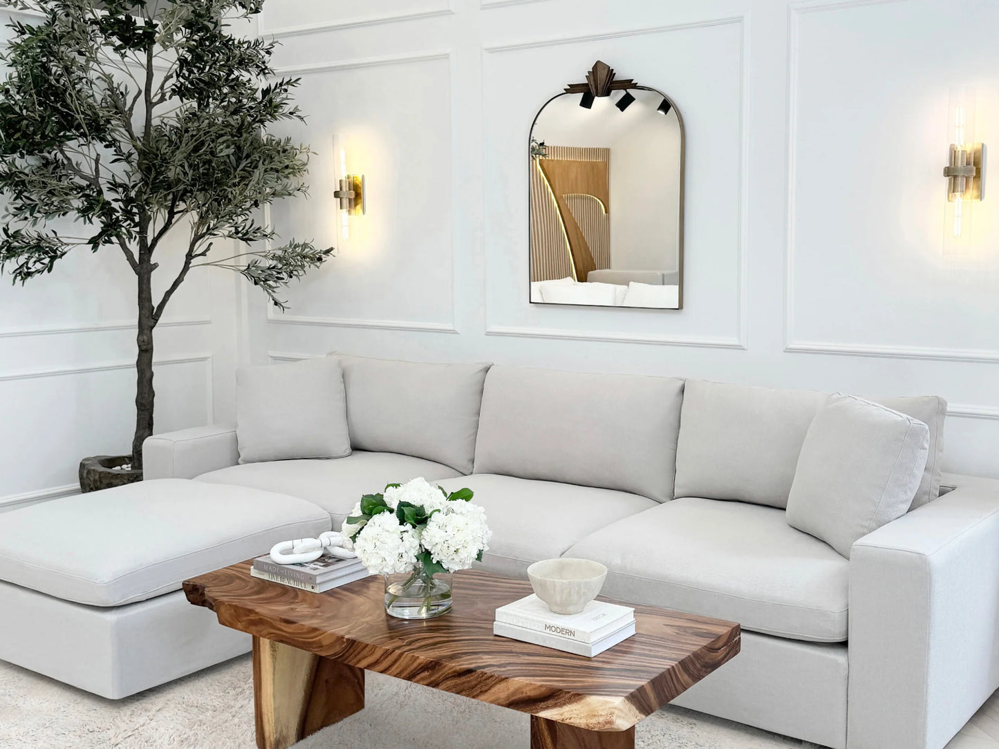 A stylish living room features a 4-Seat Modular Chaise Sectional with Ottoman in white, accompanied by a wooden coffee table adorned with flowers and decor. A large wall mirror and two wall sconces enhance the space, while a potted tree is placed in the corner against a paneled white wall.