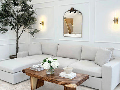 A stylish living room features a 4-Seat Modular Chaise Sectional with Ottoman in white, accompanied by a wooden coffee table adorned with flowers and decor. A large wall mirror and two wall sconces enhance the space, while a potted tree is placed in the corner against a paneled white wall.