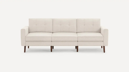 The Nomad Fabric Sofa is a light gray, three-seater with tufted back cushions, wooden legs, and a minimalist design, set against a plain white background.