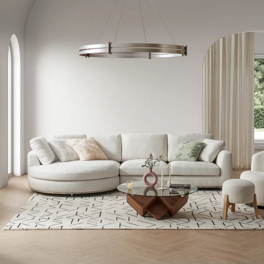 A modern living room features a white curved sectional sofa with assorted cushions, a geometric wood and glass coffee table, and a large circular chandelier. A patterned rug covers the wooden floor. Arched windows with curtains reveal greenery outside.