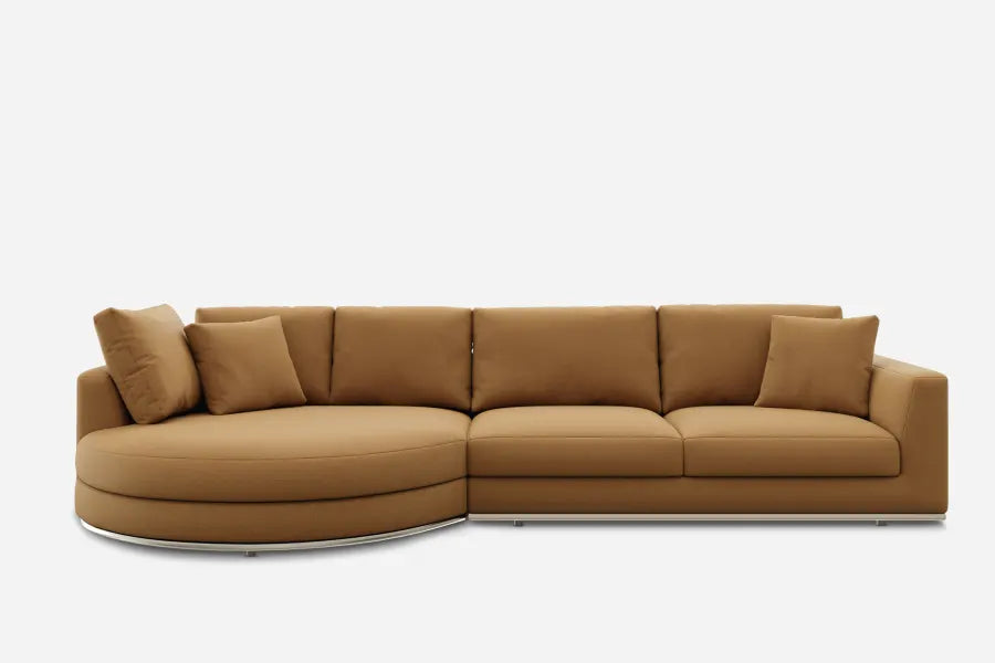 A large, modern sectional sofa in a light brown color. The left side features a rounded chaise, and it includes plush cushions. The sofa has a minimalist design and rests on a slim, metal base against a white background.