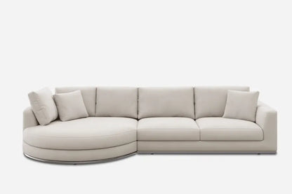 A modern beige sectional sofa with a round chaise on the left and three rectangular seat cushions. It has a clean, minimalist design and includes several matching throw pillows. The sofa is set against a plain white background.