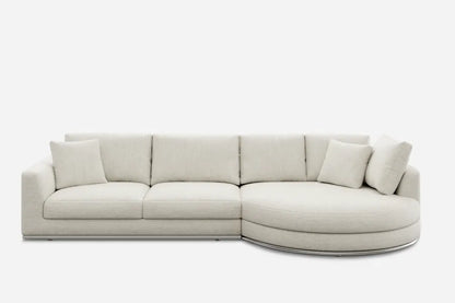 A modern, light beige sectional sofa with a rectangular design and a round chaise on the right end. It includes several matching cushions and is set against a plain white background.