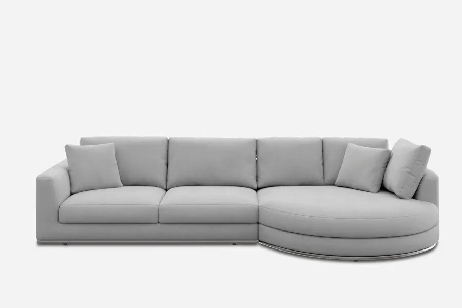 A modern, light gray sectional sofa with a clean design. It features a long, linear back with four seat cushions, complemented by a round chaise on the right side and two matching throw pillows. The sofa rests on a minimalist base.