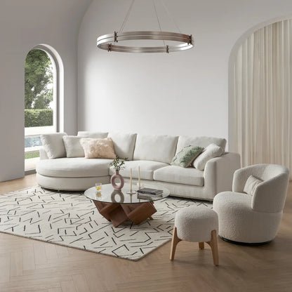 A modern living room with a white sectional sofa, a round glass coffee table, a plush armchair, and a matching ottoman. A geometric rug covers the wooden floor, and a circular chandelier hangs above. Large arched window offers a view of greenery outside.