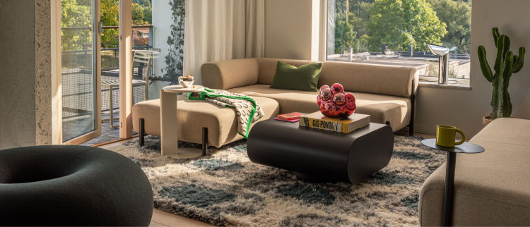 A modern living room with a beige sofa and a unique cushioned armchair, both accented with green pillows. Two wooden nesting coffee tables hold several books and a bowl. An ottoman sits adjacent to the tables, and the room is bathed in natural light from large windows.