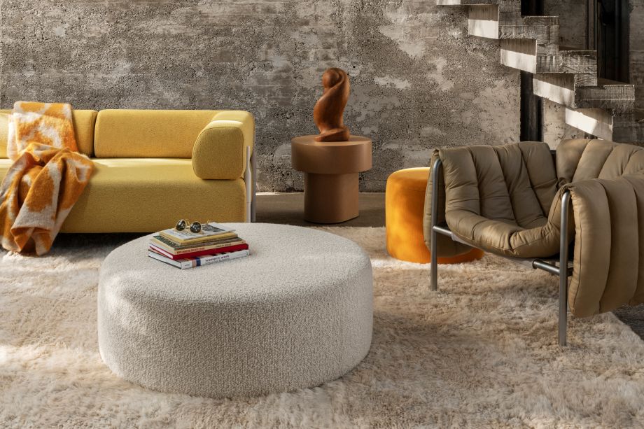 A modern living room featuring a mustard yellow sectional sofa with a white ottoman coffee table in front. A cozy rug covers the floor, and a wooden sculpture sits on a side table. Large windows provide natural light, with a concrete staircase in the background.