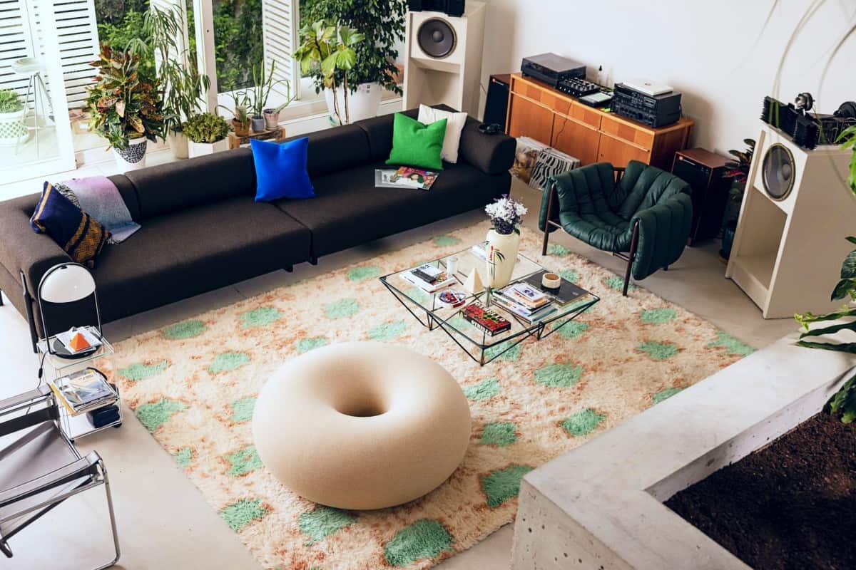 A cozy, well-lit living room featuring a dark sectional sofa adorned with colorful cushions, a unique beige donut-shaped ottoman, a glass coffee table with books and decor, a green accent chair, various plants, and vintage stereo equipment arranged in the background.