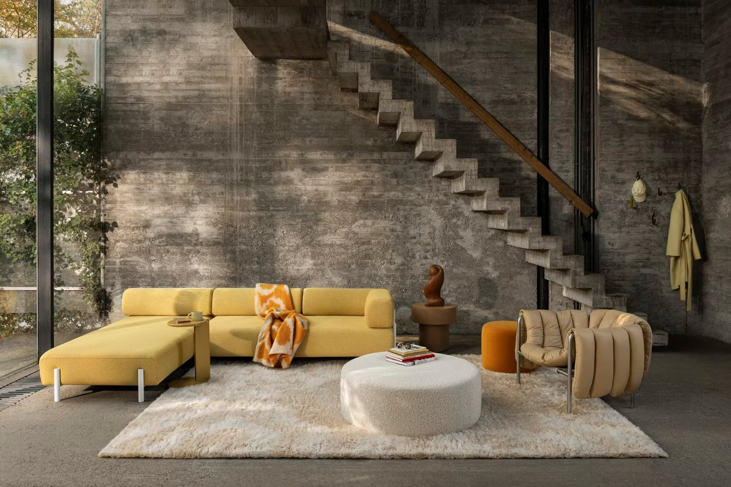 A modern living room featuring a yellow sectional sofa with a throw blanket, a beige round ottoman, a light brown armchair, and a small side table with a decorative item and books. The room has a concrete wall with a staircase and a tall window showing greenery outside.