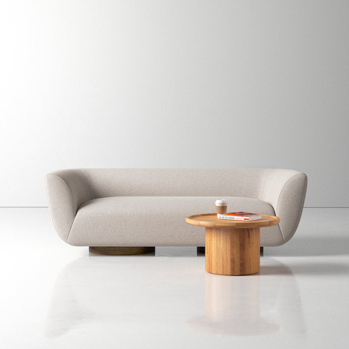 A minimalist living room features the Holloman 87'' Upholstered Sofa in light gray, situated on a glossy white floor. A round wooden coffee table placed in front of the sofa holds a coffee cup and an open magazine, against a plain, light-colored background wall.