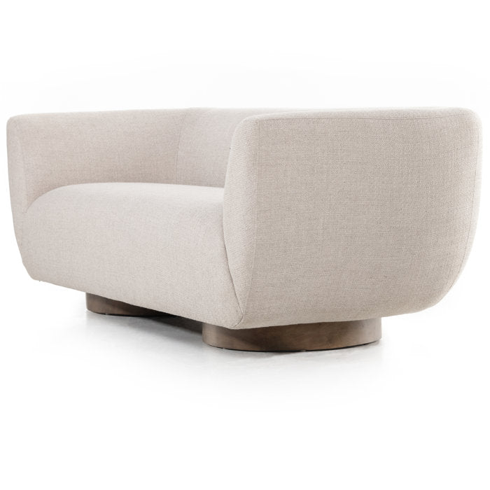 The Holloman 87'' Upholstered Sofa is a contemporary beige piece with a curved back and arms. It features smooth, plush fabric and is supported by a sturdy wooden base, elegantly showcased against a white background.