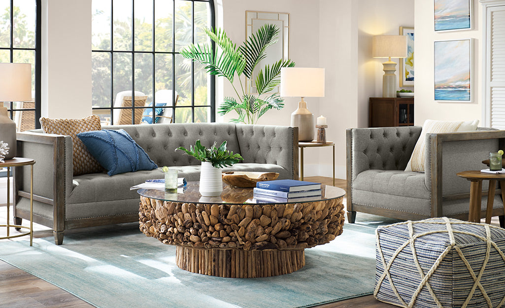 A modern living room with a curved white sofa adorned with a black pillow and a beige throw blanket. A round wooden coffee table and a side table with plants and books sit on a patterned rug. An arched floor lamp and a large plant decoratively complete the space.