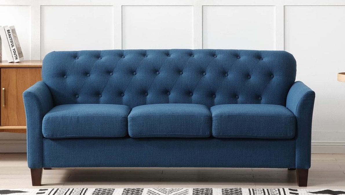 A blue tufted three-seater sofa with dark wooden legs is placed against a white paneled wall. To the left is a wooden cabinet with a couple of books on top. A portion of a patterned black and white rug is visible on the floor.