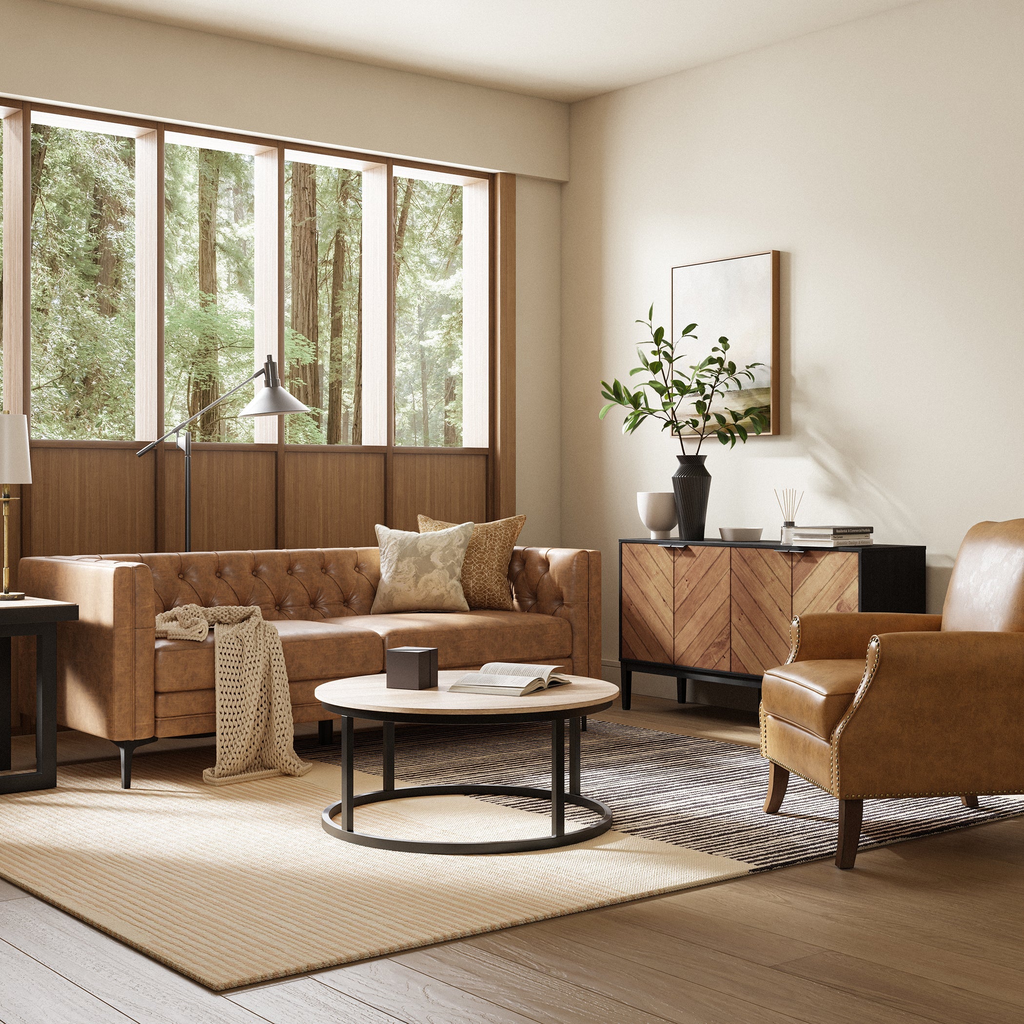 A cozy living room featuring a tan leather sofa, matching armchair, and a round wooden coffee table. A woven rug and large windows showcasing a lush green outdoor view enhance the space. A wooden sideboard with decorative items completes the inviting atmosphere.