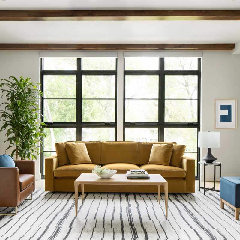 A modern living room features a navy-blue sofa set with white and gray cushions. Three wall mirrors hang above the sofas on dark blue walls. Potted plants and a white coffee table with plants on it are placed on a white fluffy rug, adjacent to a large window with white curtains.