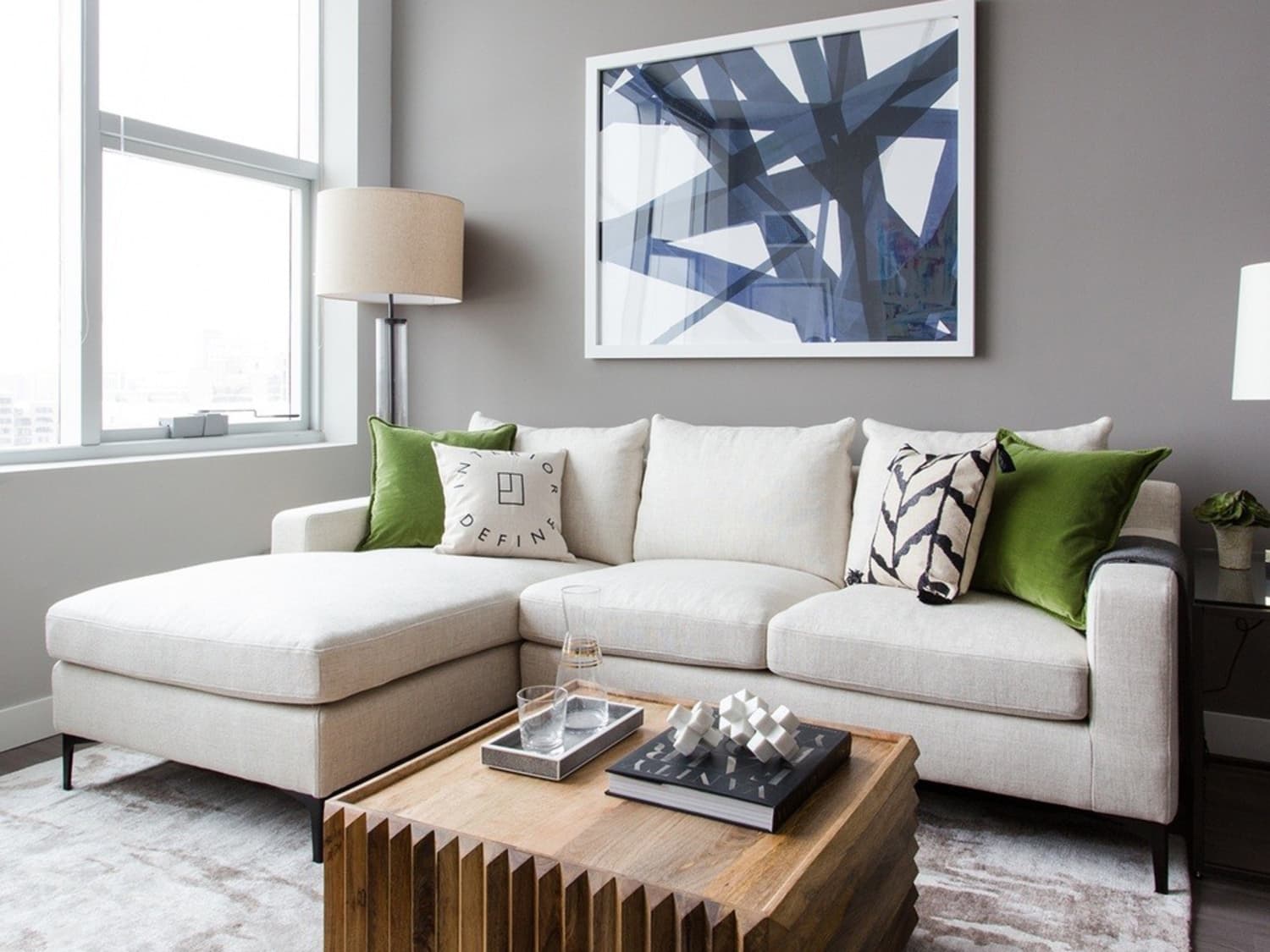 A modern living room featuring a grey sofa adorned with blue and grey cushions, a light grey coffee table with a white vase and book, and a side table with a white lamp. The walls are decorated with two framed circular art pieces. A large window brightens the space.