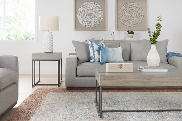 A modern living room features a dark gray sectional sofa with a plaid pillow and a throw blanket. A framed abstract artwork hangs above the sofa. A round wooden coffee table with decorative items sits in front. A large plant and side table with a lamp are in the background.