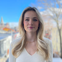 Woman with long blonde hair wearing a white top stands outdoors. She has a neutral expression, and the background features a clear blue sky and blurred urban landscape with buildings and trees.