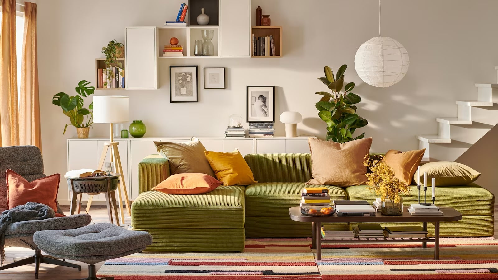 A cozy living room featuring a green sofa with an assortment of colorful cushions. A modern lounge chair and footrest sit nearby. The room is adorned with potted plants, a bookshelf, framed pictures, a white pendant light, and a multicolored striped rug.