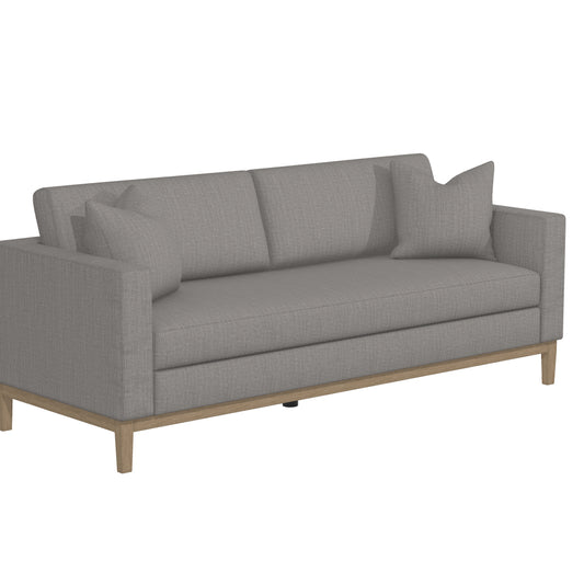 The Capri Sofa is a modern piece with clean lines and wooden legs, finished in a light gray color. It includes two cushions and two throw pillows, offering a simple yet elegant design perfect for a contemporary living space.