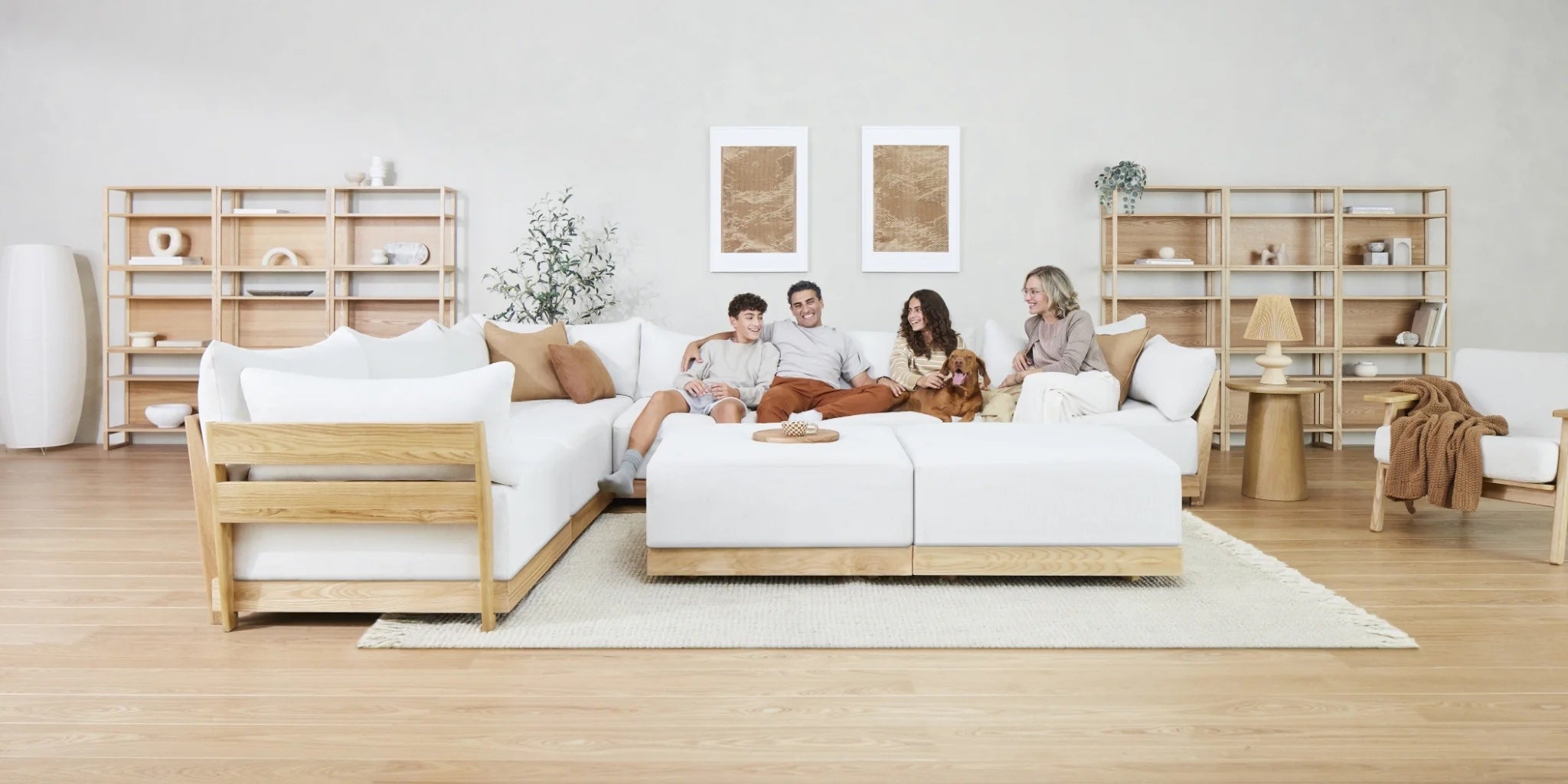 A family of four sits on a modern white sectional sofa with a dog, facing the camera and smiling. Behind them are wooden shelves filled with decor items. The room has a minimalistic design with neutral tones, a hardwood floor, and framed artwork on the wall.