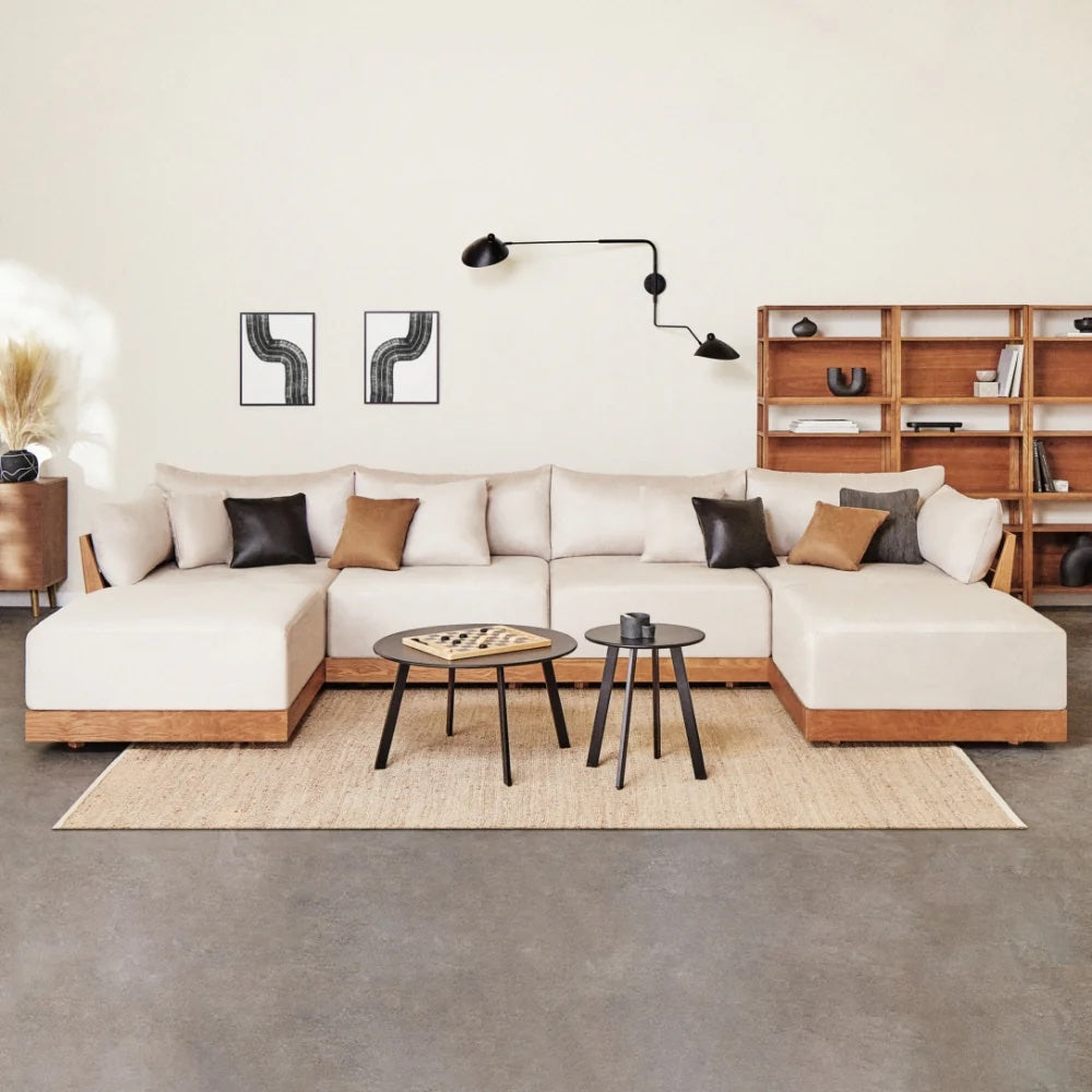 A modern living room with an L-shaped beige sectional sofa adorned with black and brown cushions. The room features a wooden bookshelf, two abstract art pieces on the wall, a unique black wall lamp, and a set of round coffee tables on a light rug.