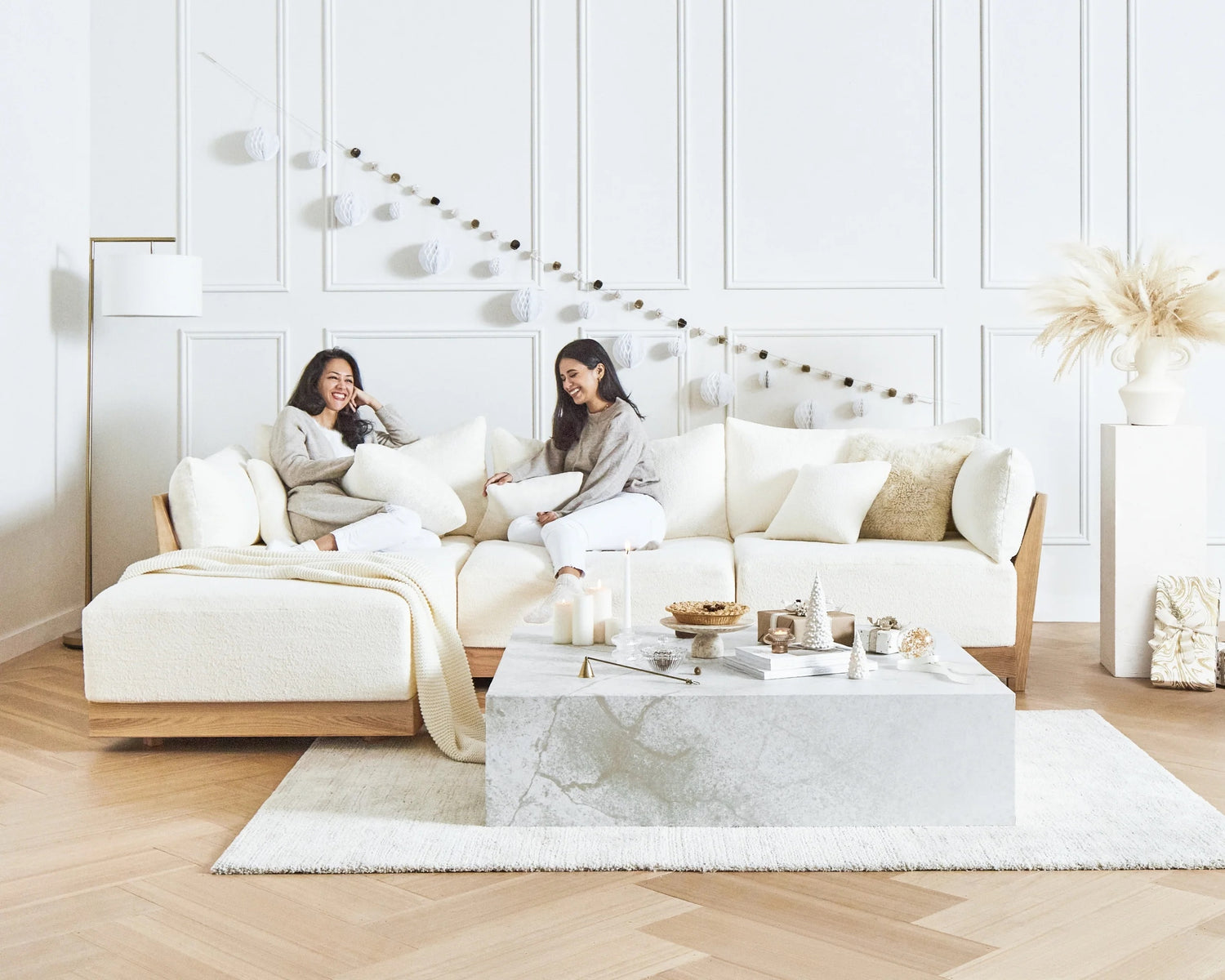 A serene living room features two women sitting and laughing on a cozy, cream-colored sectional sofa adorned with pillows and a throw. The decor includes a marble coffee table with snacks and candles, pampas grass in a vase, and string lights on a paneled white wall.