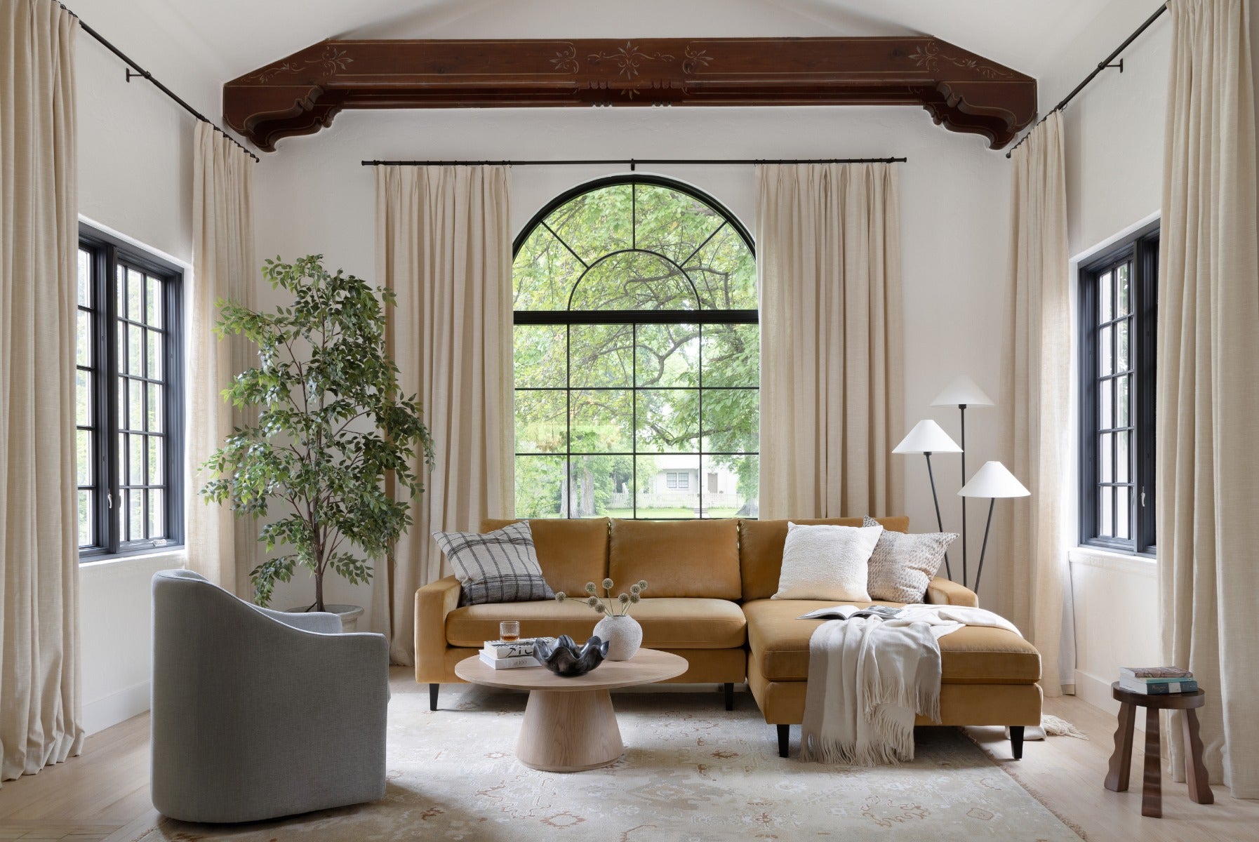 A cozy living room features a mustard-colored sectional sofa adorned with throw pillows and a blanket. A round wooden coffee table holds decor items. A large arched window with black frames and beige curtains opens to greenery outside. A floor lamp and an accent chair complete the space.