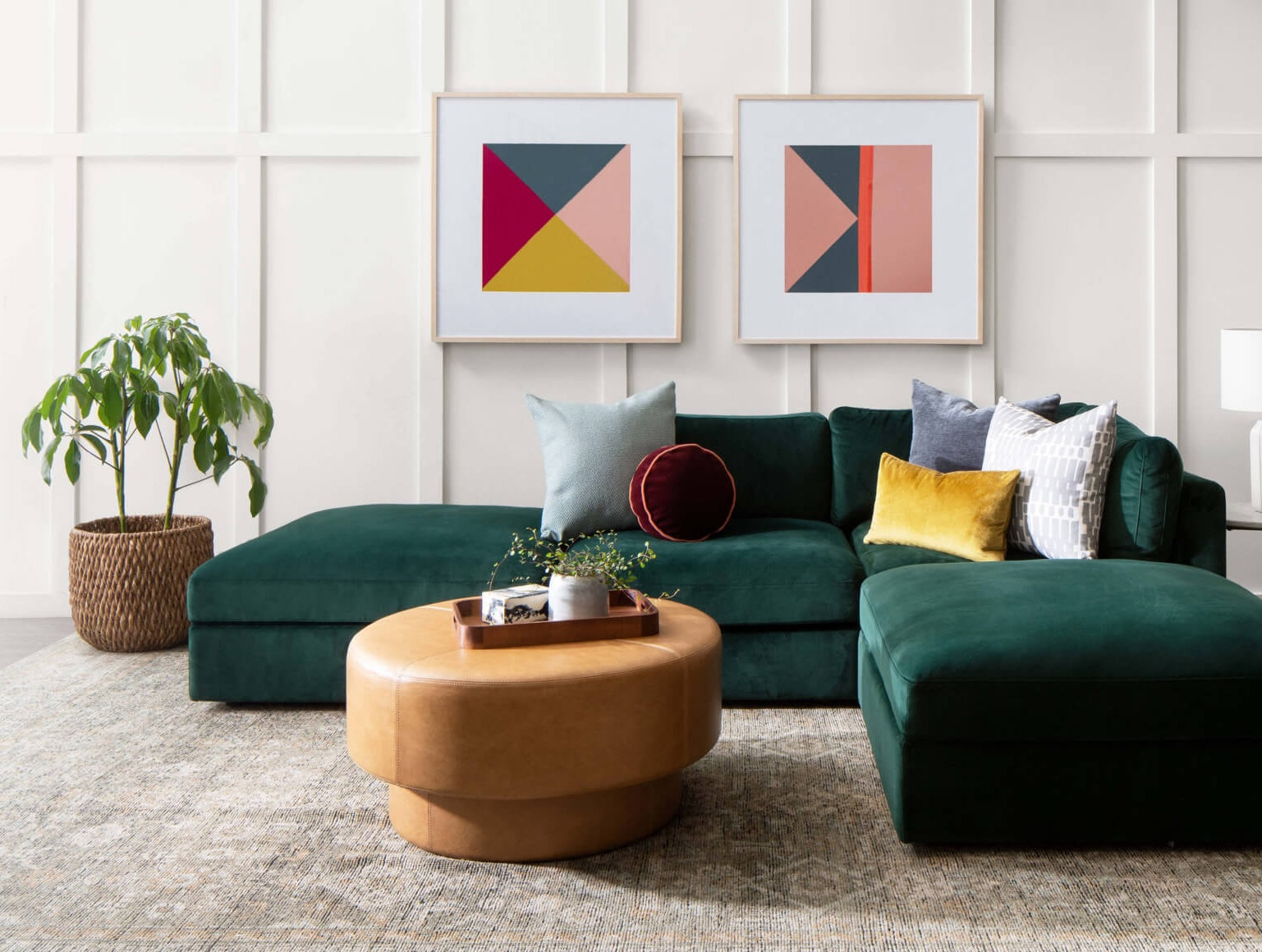 A modern living room features a dark green sectional sofa adorned with assorted cushions, a brown round coffee table with a plant on top, and two colorful geometric prints on the wall. A potted plant sits to the left, and the room has a patterned neutral rug.