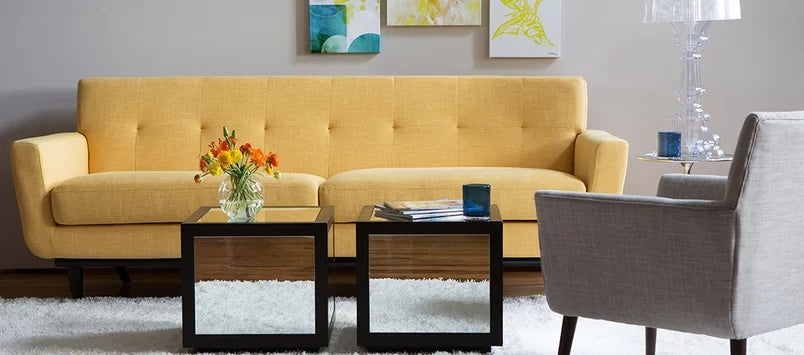 Living room with a modern design featuring a yellow sofa, two black square coffee tables with glass tops, and a grey armchair. A vase with orange flowers and books decorate the tables. Colorful wall art and a clear lamp add accents to the space with a white rug.