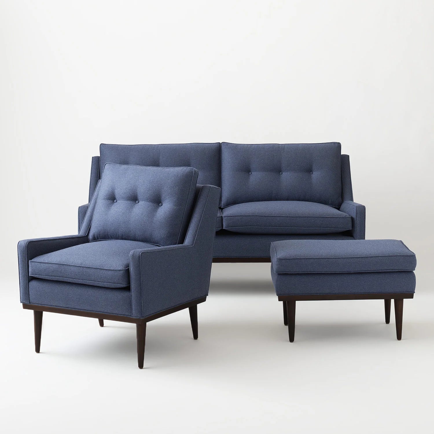 The Jack Sofa collection includes a modern living room set featuring a dark blue fabric sofa, armchair, and ottoman. Each piece boasts wooden legs and a minimalist design, beautifully arranged on a light background.