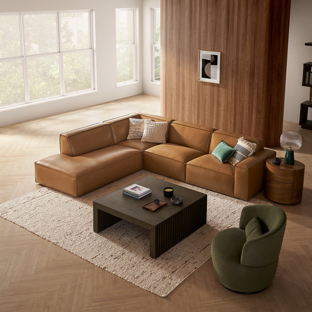 A modern living room showcasing the Jonathan Leather Extended Side Chaise Sectional in a tan finish, embellished with cushions. The space also includes a dark green swivel chair, a textured rug, and a rectangular coffee table. Large windows illuminate the area, which features a wooden accent wall adorned with framed artwork.