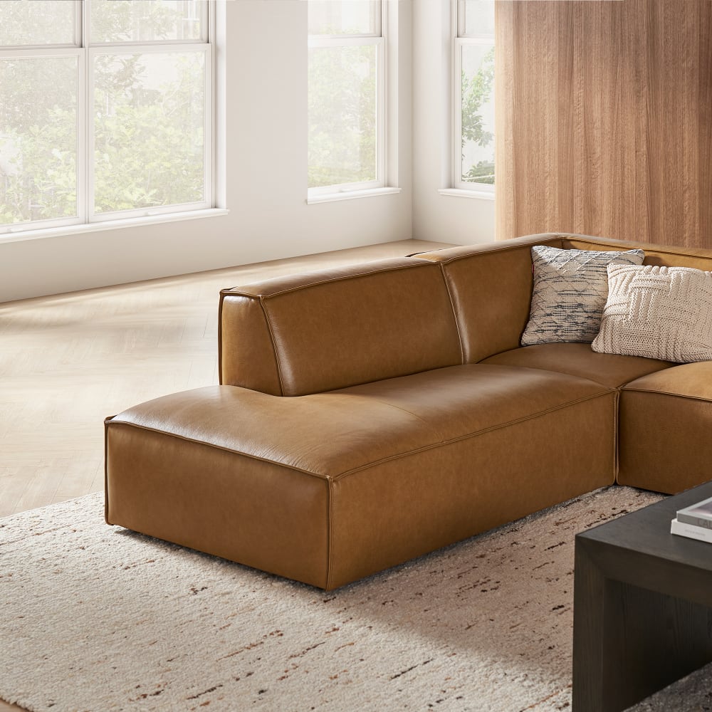 A modern living room features the Jonathan Leather Extended Side Chaise Sectional in brown on a textured rug, with several cushions thoughtfully arranged on the sofa. Large windows let in natural light, while a wooden wall adds warmth to the space.