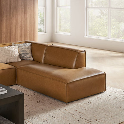 A modern living room showcases the Jonathan Leather Extended Side Chaise Sectional in brown, adorned with patterned pillows. Sunlight filters through large windows, harmonizing with the light wood flooring and a neutral-toned rug, creating a minimalist and cozy atmosphere.