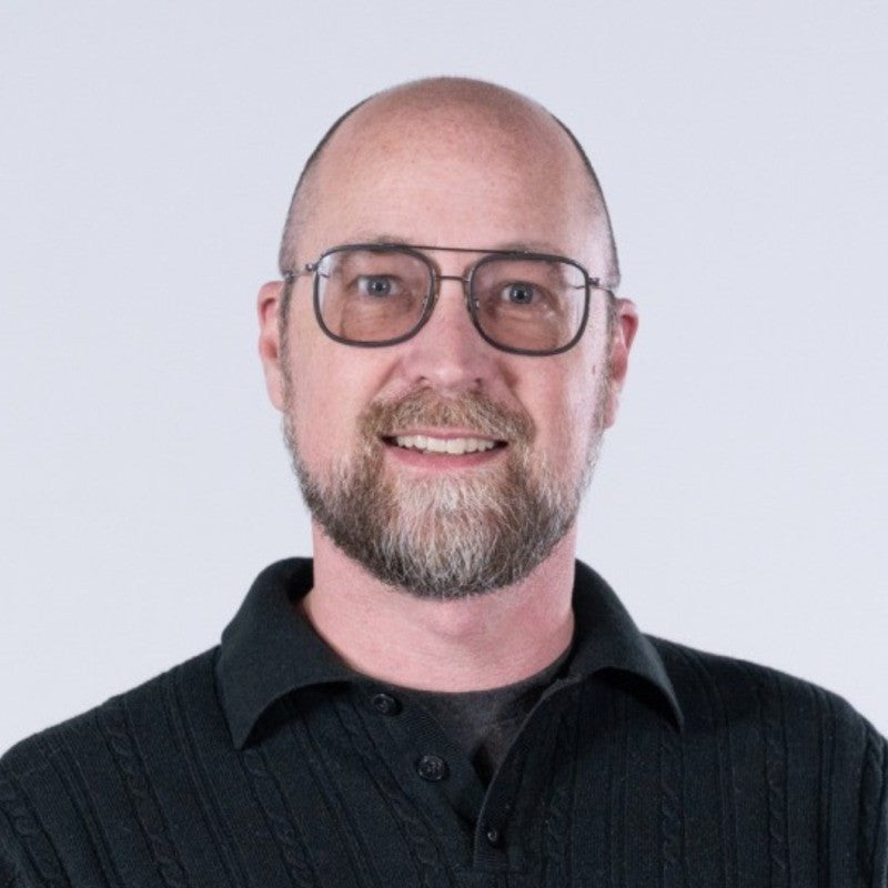 A bald man with a short beard and mustache is wearing glasses and a black sweater. He is smiling and posing against a white background.