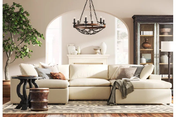 A cozy living room with a cream sectional sofa adorned with throw pillows and a blanket. A round chandelier hangs from the ceiling. The room features a side table with a vase, a large indoor plant, a decorative cabinet, and a patterned rug on wooden flooring.