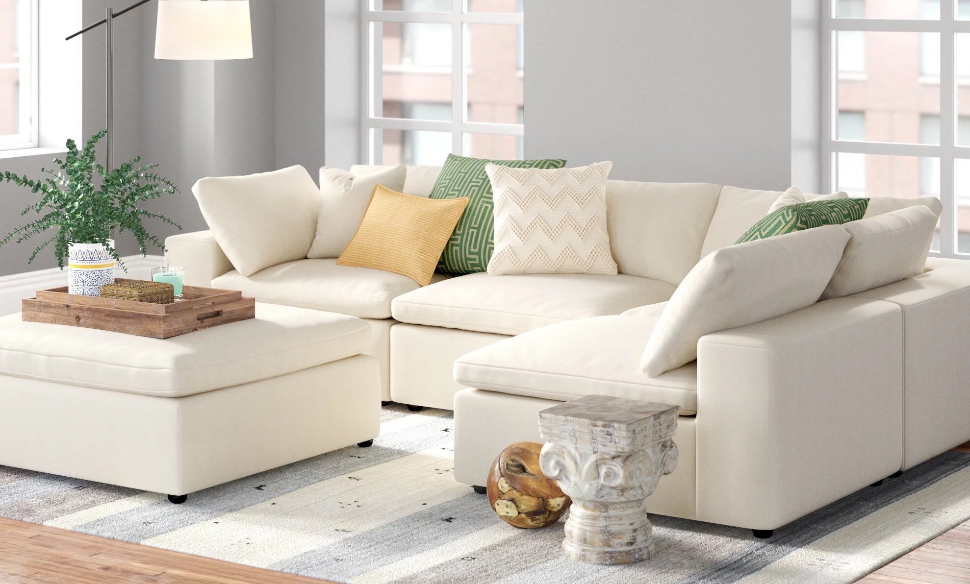 A modern living room with a large, white sectional sofa adorned with colorful throw pillows, including shades of yellow and green. A wooden coffee tray with decorative items sits on an ottoman, while a small decorative stone side table and indoor plant are nearby.