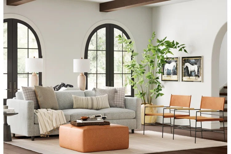 A bright, modern living room features a gray sofa with assorted pillows, a brown leather pouf, two brown leather chairs, and a black coffee table. Tall, arched windows, a potted plant, framed horse artwork, and white walls complete the space.