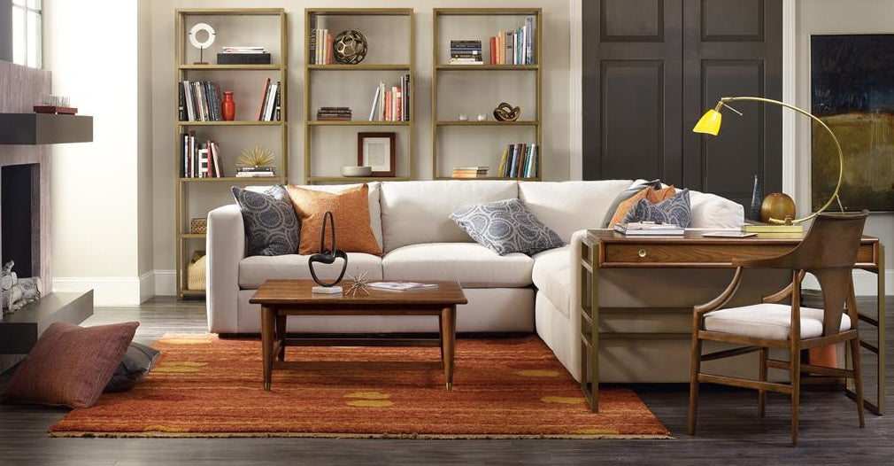 A cozy living room with a beige sectional sofa adorned with patterned cushions. A wooden coffee table sits on a red-orange rug. Behind the sofa is a wooden desk with a chair and a gold desk lamp. Bookshelves with books and decor stand against the wall.