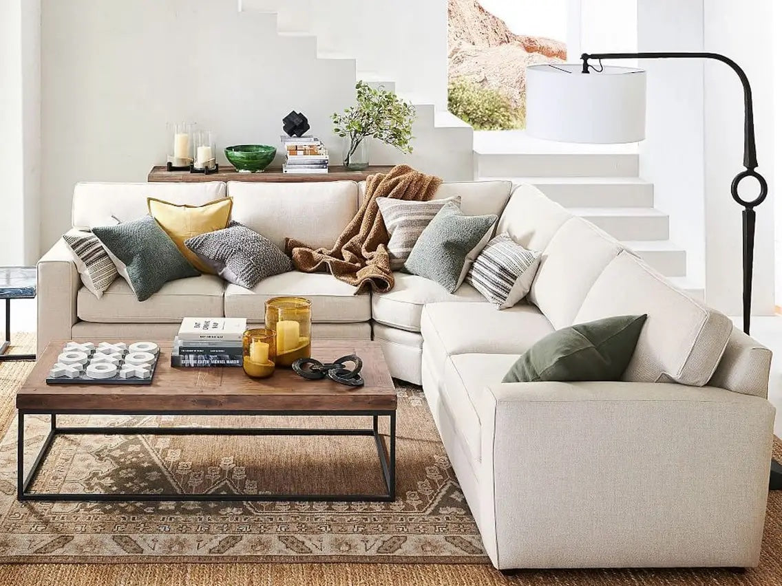 A cozy living room featuring a L-shaped beige sofa with various pillows and a throw blanket. A wooden coffee table with candles, books, and decorative items sits on a patterned rug. The background shows white stairs leading up to a window with natural light.