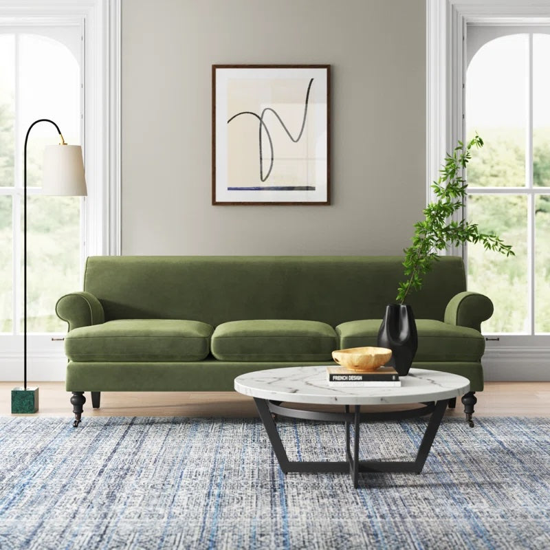 A modern living room features a green velvet sofa with rolled arms, placed against a light gray wall. Above it hangs an abstract framed art piece. A round coffee table with a white top and black legs holds a vase with greenery and a decorative bowl. A floor lamp stands to the left. The room has large windows and a blue patterned rug.