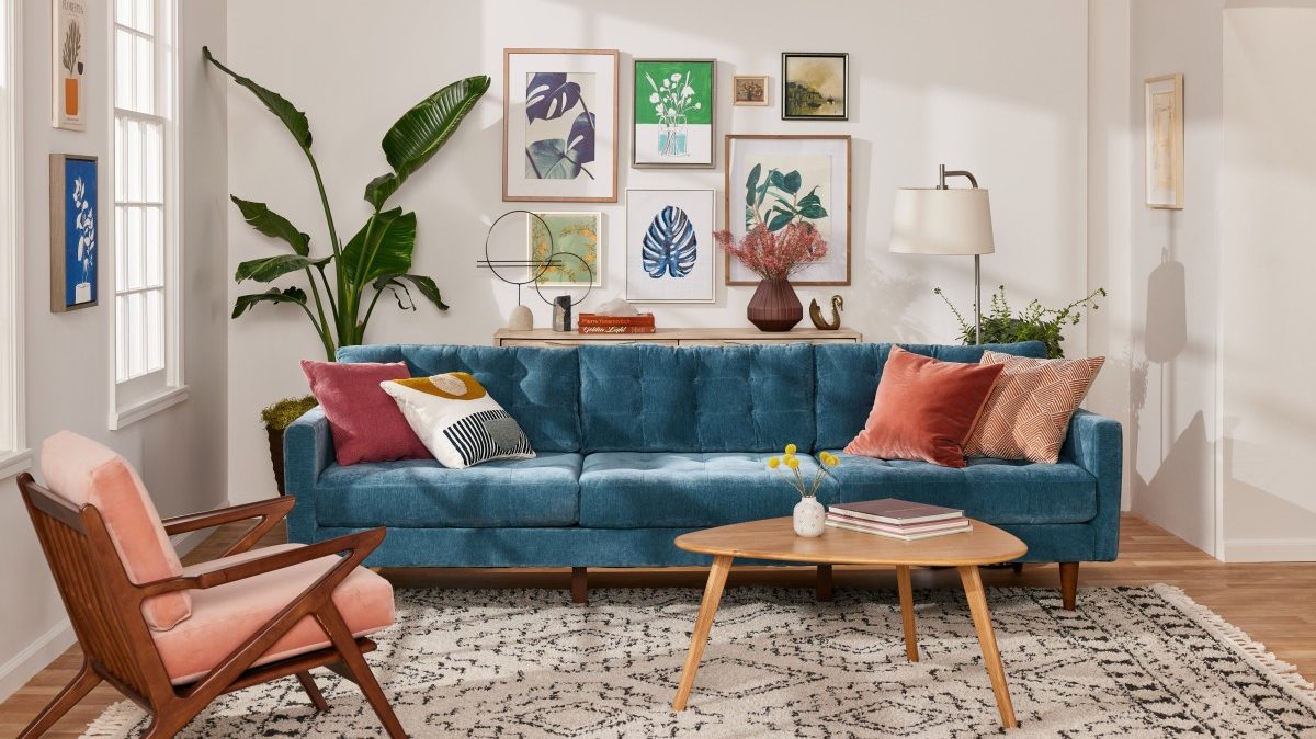 A modern living room features a blue tufted sofa adorned with colorful pillows, a mid-century wooden coffee table, and a coral chair. Artwork and plants decorate the white walls, and a large window brightens the space. A cozy rug completes the room.