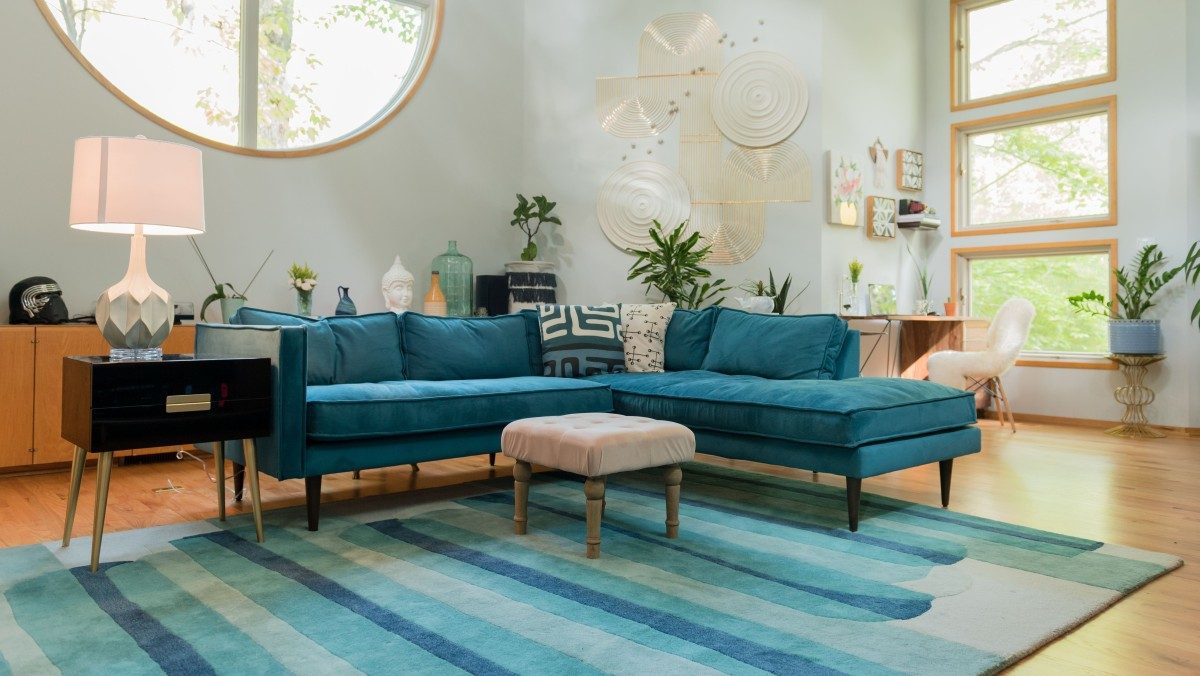 A modern living room with a teal sectional sofa, a light pink ottoman, and a striped blue area rug. Decor includes wall art, plants, and various decorative objects. Large round and rectangular windows allow natural light to fill the room, highlighting wooden flooring.