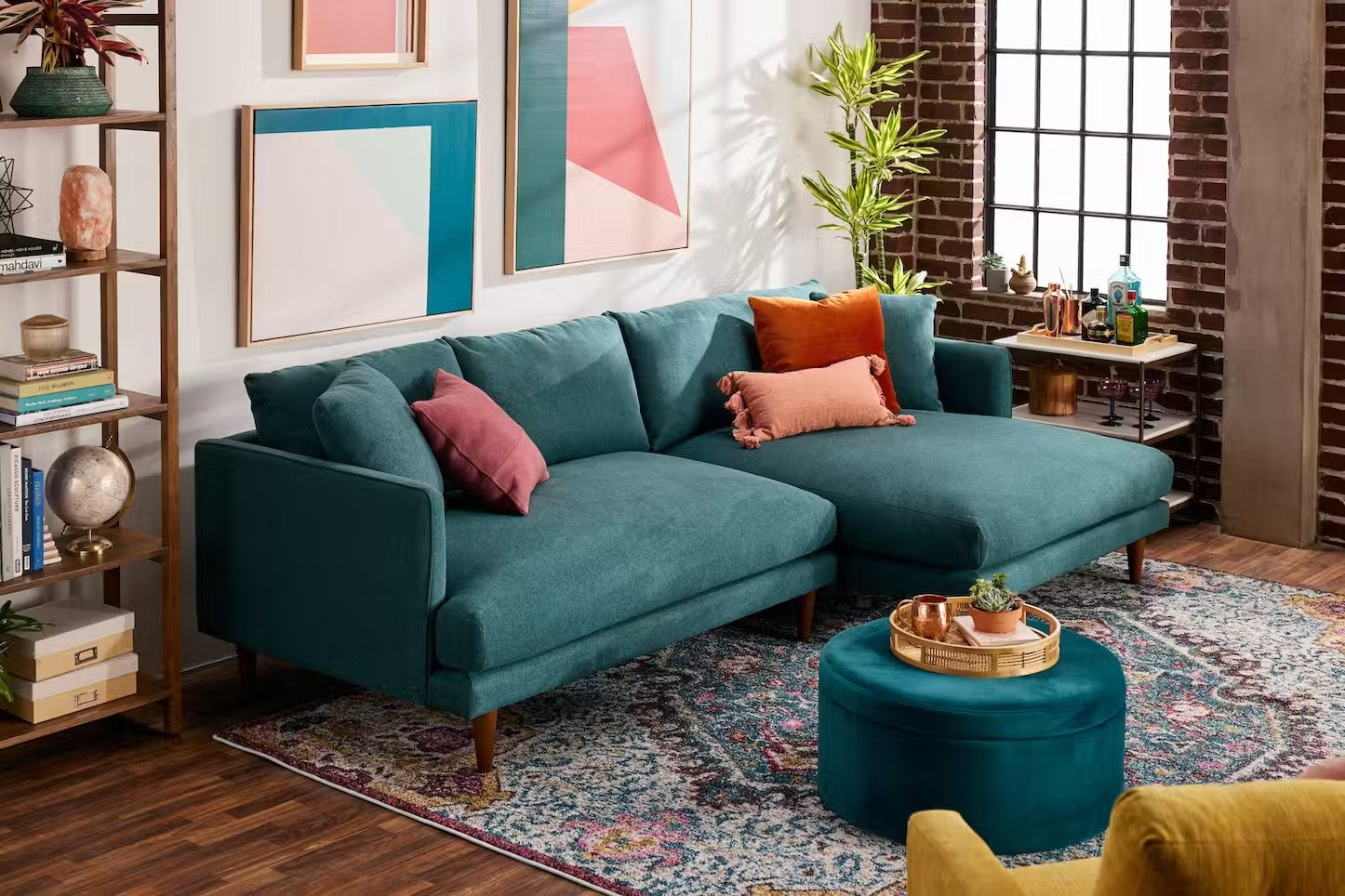 A stylish living room features a teal sectional sofa adorned with colorful cushions and a matching round ottoman. Abstract art pieces decorate the walls, and a large window allows natural light to fill the space. A rug accents the wooden floor and a plant adds greenery.