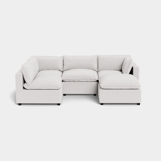 The Kova L Shape with Ottoman is a contemporary, modular light gray sofa featuring clean lines and plush cushions in an L-shaped design. It showcases square armrests and rests on a minimalist black base, set against a plain white background.