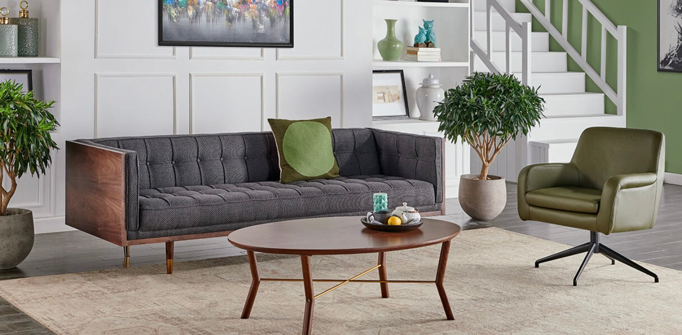 A modern living room features a gray tufted sofa with a green throw pillow, a round wooden coffee table with decor items, and a green upholstered chair. The background includes shelves with decorative items, a staircase, and a mix of white and green walls.