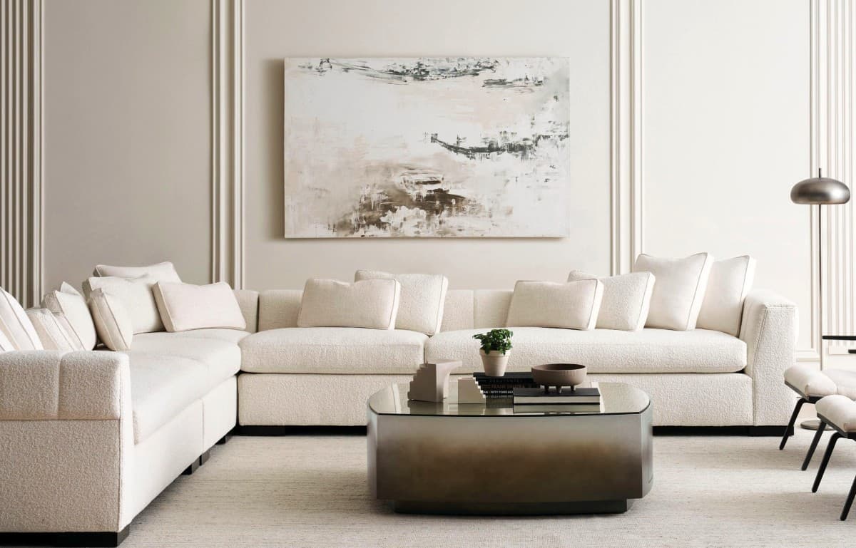 A modern living room features an L-shaped white sectional sofa with numerous cushions. A minimalist abstract painting hangs on the wall above. In front of the sofa is a glass-topped coffee table with books and a small plant. The decor is neutral and contemporary.
