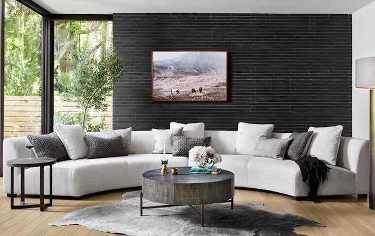 A modern living room featuring a curved white sectional sofa with numerous gray and white pillows. A round coffee table sits on a gray rug. The wall behind the sofa is black with a framed landscape photograph. Floor-to-ceiling windows reveal a lush outdoor scene.