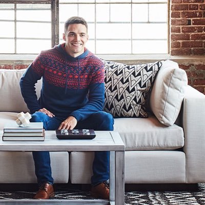 A smiling person in a blue and red patterned sweater sits on the edge of a light-colored couch, with a patterned pillow and books on the coffee table in front. The room features exposed brick, large windows, and modern decor.
