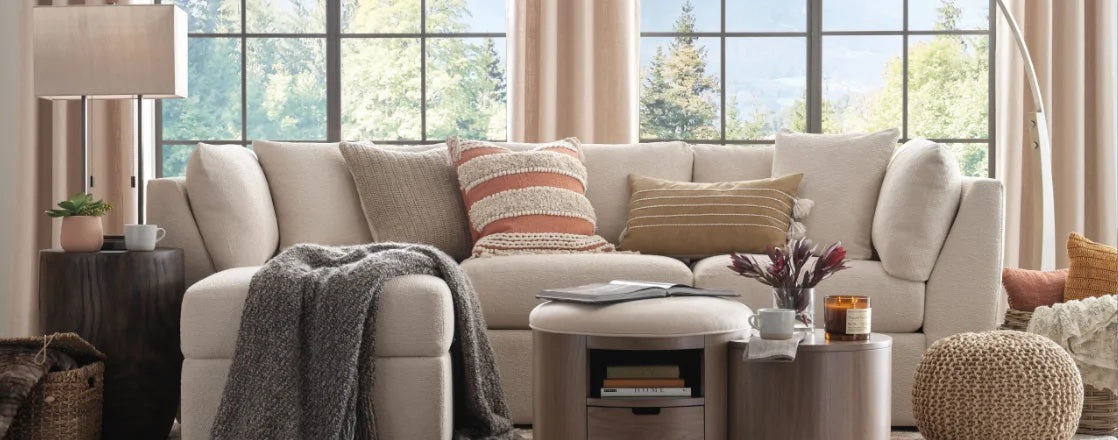 A cozy living room featuring a cream-colored sectional sofa with assorted patterned throw pillows. A gray knit blanket is draped over the sofa. The room includes a wooden side table with a lamp, a round ottoman, and large windows with views of trees and mountains.